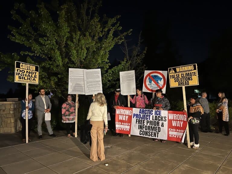 Indigenous protestors hold signs saying Danger Do Not Inhale Silica in front of a blond woman attendinga fancy event